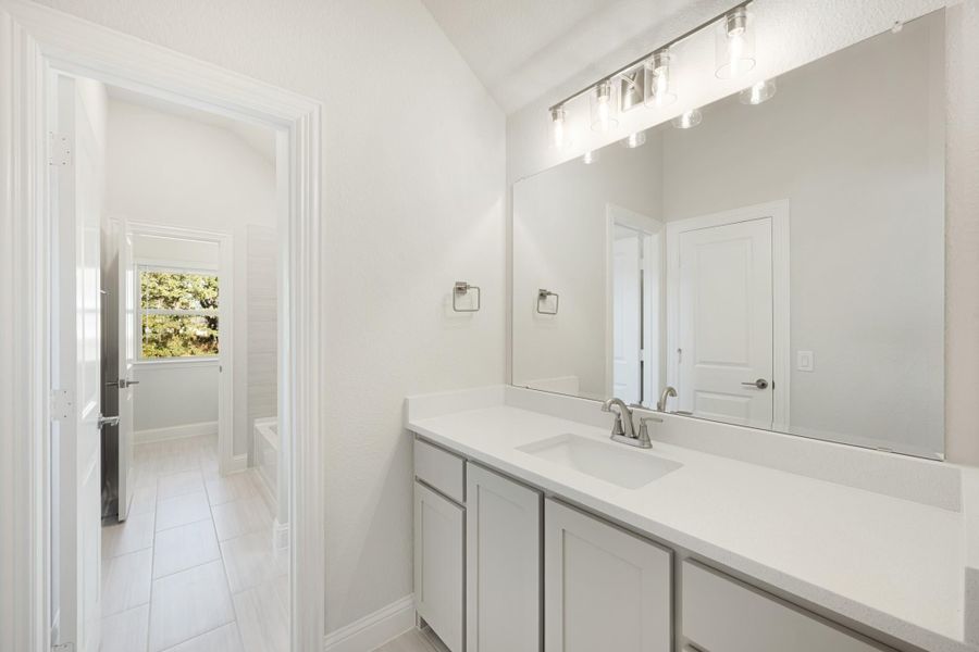 Bathroom vanity with white quartz countertop, gray cabinets, large mirror, and soaking tub visible in background