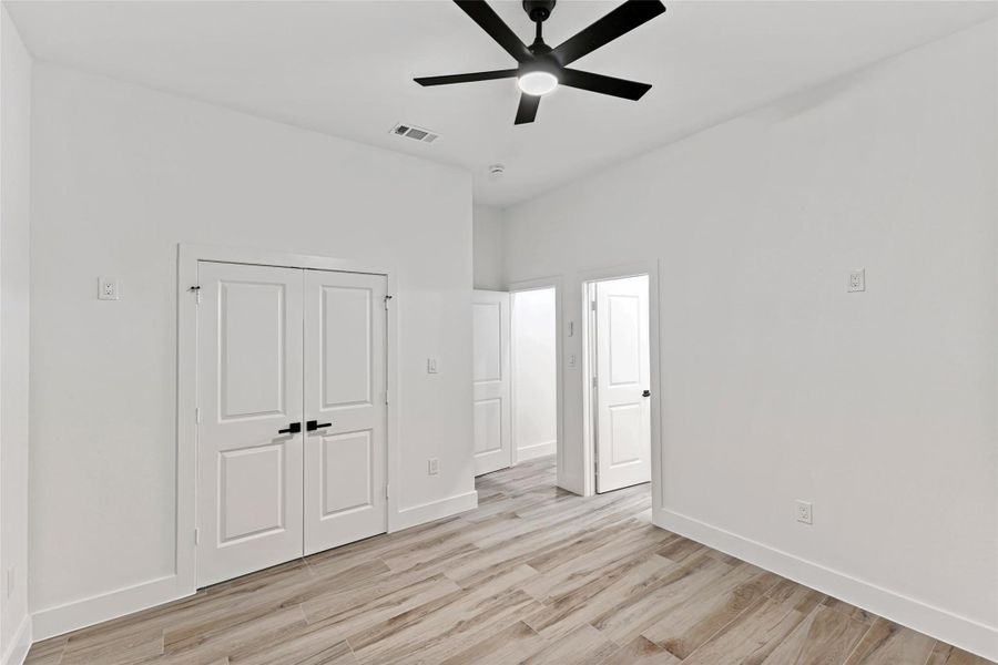 This is a bright, modern bedroom with light wood-like tile flooring, white walls, and a ceiling fan. It features a double-door closet and an additional door leading to it's own ensuite. This is a bright, modern bedroom with light wood-like tile flooring, white walls, and a ceiling fan. It features a double-door closet and an additional door leading to it's own ensuite.