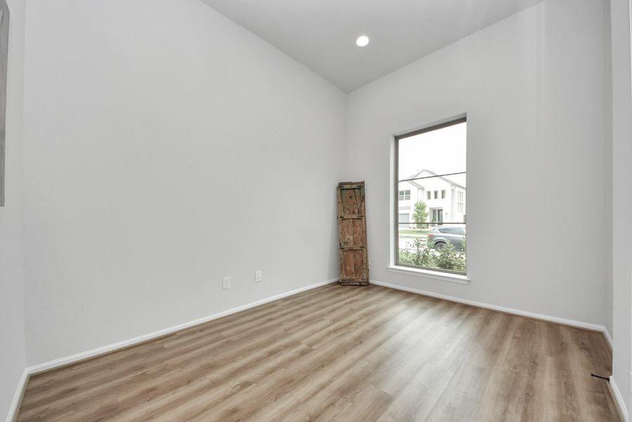 This room features a high ceiling, large window with natural light, and wood-style flooring, creating a bright and airy space perfect for an office or sitting area.