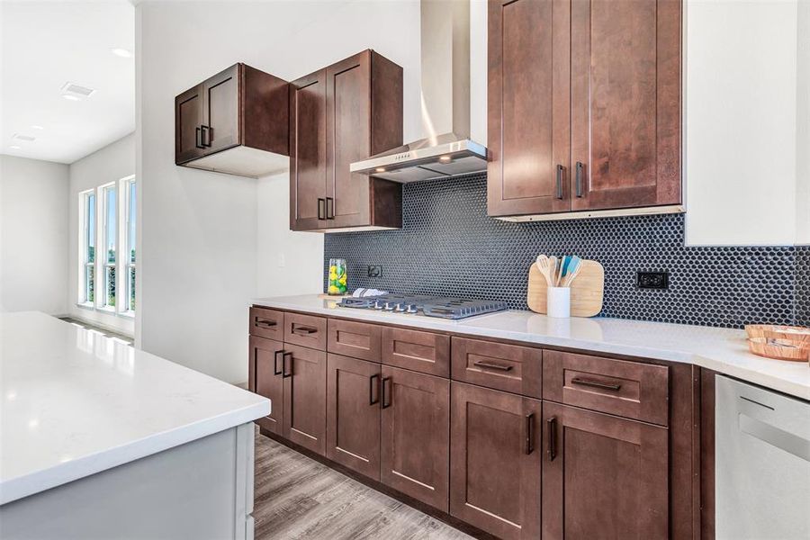 Kitchen featuring dark brown cabinets, tasteful backsplash, stainless steel gas stovetop, wall chimney exhaust hood, and light hardwood / wood-style flooring