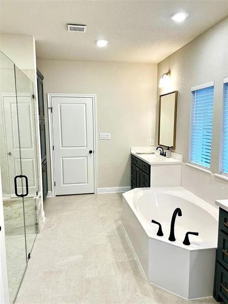 Spacious bathroom featuring a white soaking tub with black fixtures, a walk-in shower with glass enclosure, dual dark cabinetry vanities with white countertops, and light tile flooring throughout Spacious bathroom featuring a white soaking tub with black fixtures, a walk-in shower with glass enclosure, dual dark cabinetry vanities with white countertops, and light tile flooring throughout