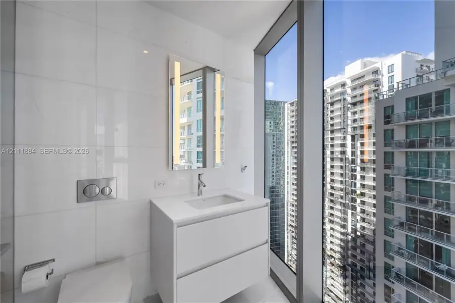 Modern floating vanity with a white countertop and rectangular undermount sink, floor-to-ceiling windows, wall-mounted dual-flush toilet, and vertical LED-integrated mirror Modern floating vanity with a white countertop and rectangular undermount sink, floor-to-ceiling windows, wall-mounted dual-flush toilet, and vertical LED-integrated mirror