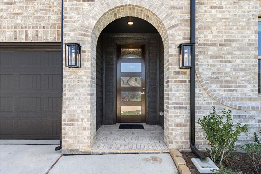 Brick home entrance with arched doorway, dark wood front door with glass panels, and wall sconce lighting