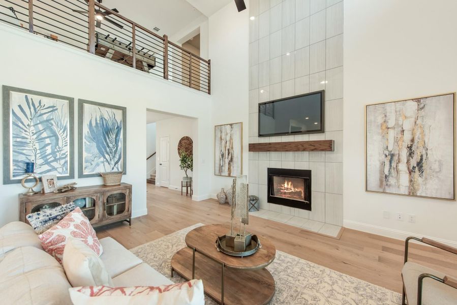 Living room with two-story ceiling, tiled fireplace, wall-mounted TV, hardwood floors, and open loft railing above. Living room with two-story ceiling, tiled fireplace, wall-mounted TV, hardwood floors, and open loft railing above.