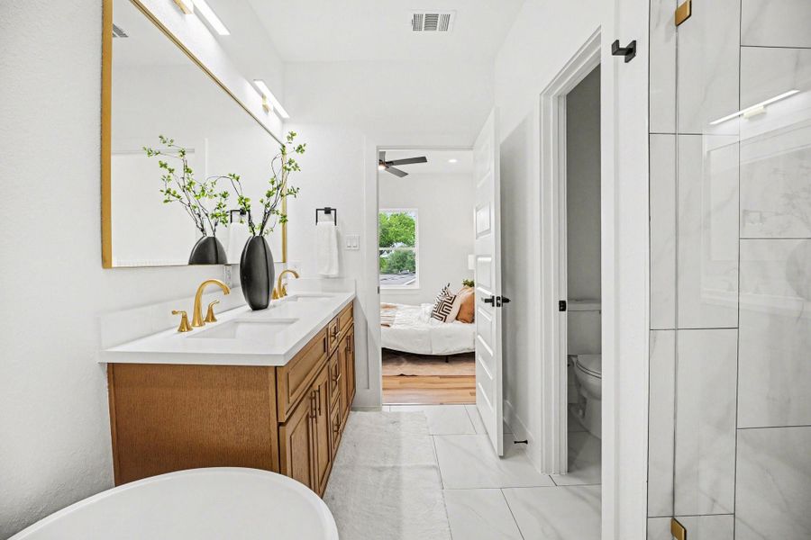 This photo showcases a modern bathroom with a spacious double vanity featuring gold fixtures, a large mirror, and decorative vases. The room is bright with white and wood accents, and there's a partial view into a cozy bedroom with a large window, hardwood floors, and neutral decor.