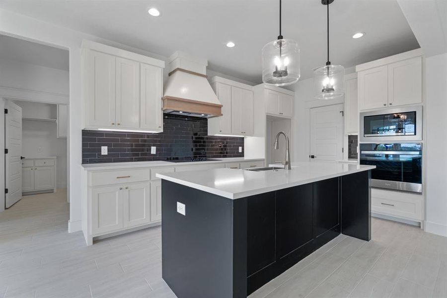 Kitchen featuring backsplash, light countertops, white cabinetry, decorative light fixtures, and custom range hood