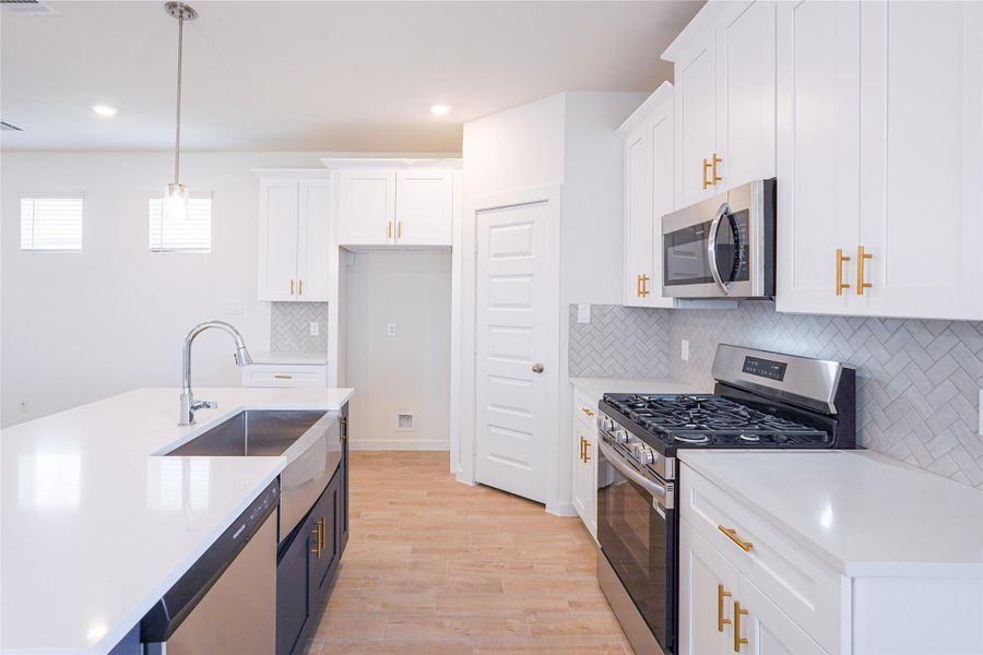 Modern kitchen featuring sleek white cabinetry with gold hardware, stainless steel appliances, and a spacious island with a dual sink. The herringbone backsplash adds a stylish touch, complemented by light wood flooring and ample natural light.