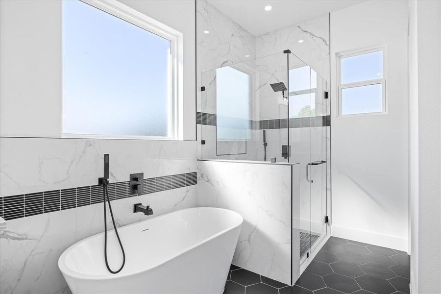 Freestanding soaking tub with matte black fixtures, marble-look tile walls with a dark accent band, frameless glass shower enclosure, and hexagonal floor tiles