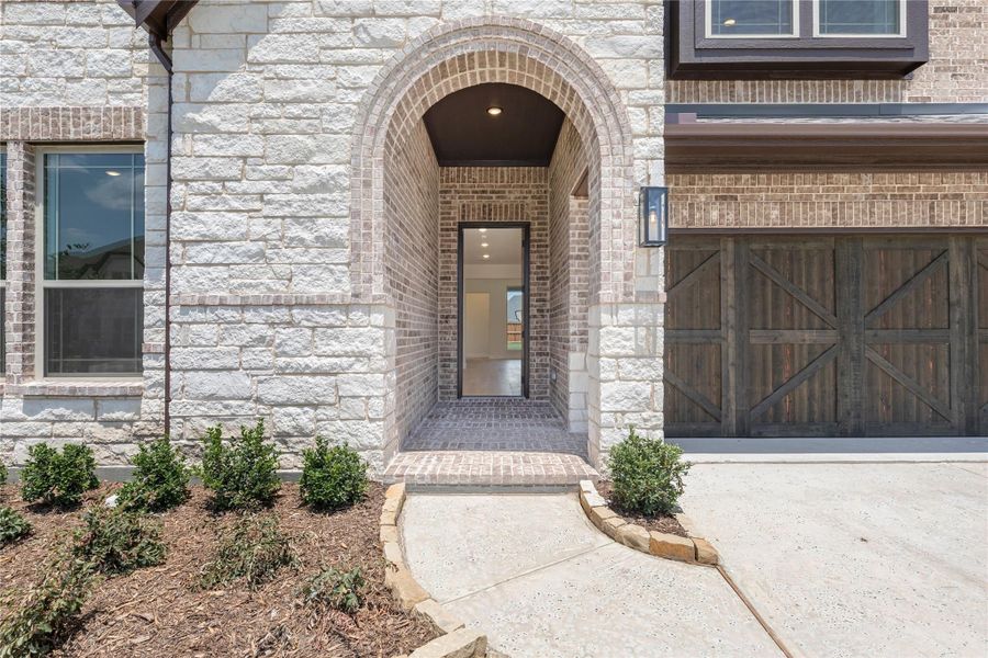 Home entrance with brick arched entryway, stone exterior, and rustic wood garage door beside landscaped shrubs