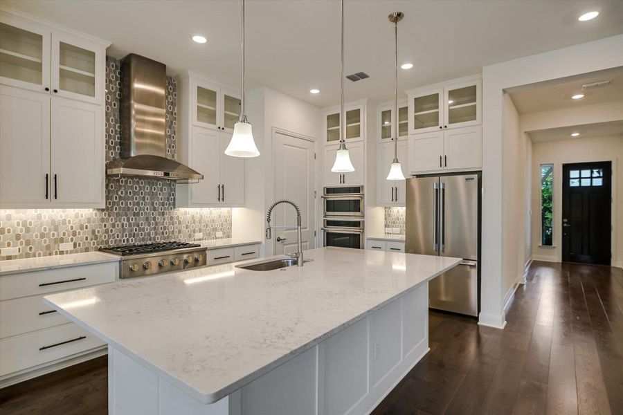 Kitchen with decorative backsplash, appliances with stainless steel finishes, wall chimney range hood, a kitchen island with sink, and dark wood-style floors Kitchen with decorative backsplash, appliances with stainless steel finishes, wall chimney range hood, a kitchen island with sink, and dark wood-style floors