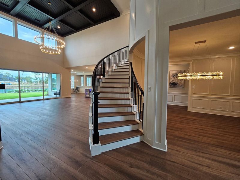 This photo showcases a spacious, elegant home interior with an open floor plan. It features a grand curved staircase, large windows providing ample natural light, and stylish chandeliers. The wood flooring and high ceilings add a touch of luxury to the modern design.