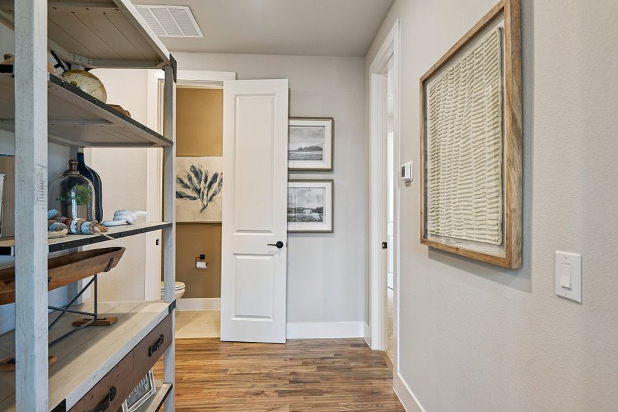 Hallway with wood floors, open shelving unit, wall art, and a half bathroom with white door