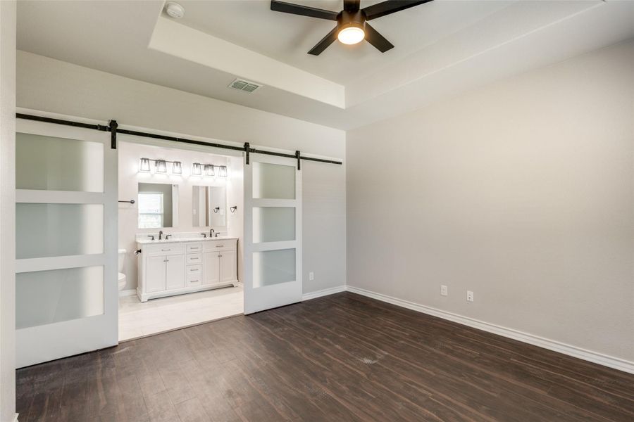 Unfurnished bedroom featuring a barn door, dark wood finished floors, a raised ceiling, ceiling fan, and ensuite bath Unfurnished bedroom featuring a barn door, dark wood finished floors, a raised ceiling, ceiling fan, and ensuite bath
