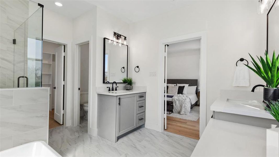 This bright bathroom features a spacious layout with a glass-enclosed shower, a modern vanity with black fixtures, and elegant tile flooring. The room connects to a bedroom through a sliding door, enhancing accessibility and flow.