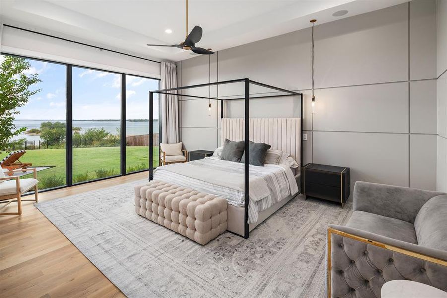 Serene primary bedroom thoughtfully designed to capture stunning lake views through expansive windows. Real wood floors, a sophisticated accent wall, and upgraded designer lighting create a luxurious retreat that blends modern elegance with the beauty of the surrounding landscape.