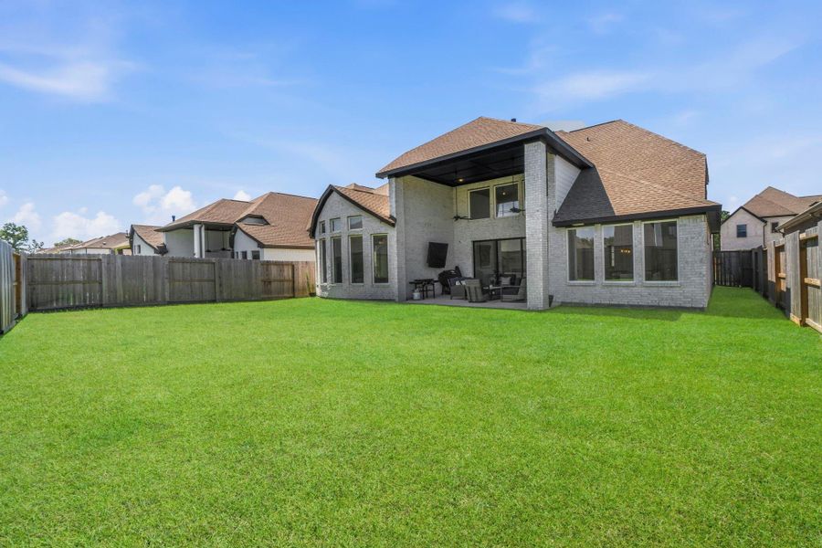 This photo shows a spacious backyard with a well-maintained lawn and a modern two-story house featuring a covered patio, ideal for outdoor entertaining. The property is fenced for privacy and surrounded by similar homes.