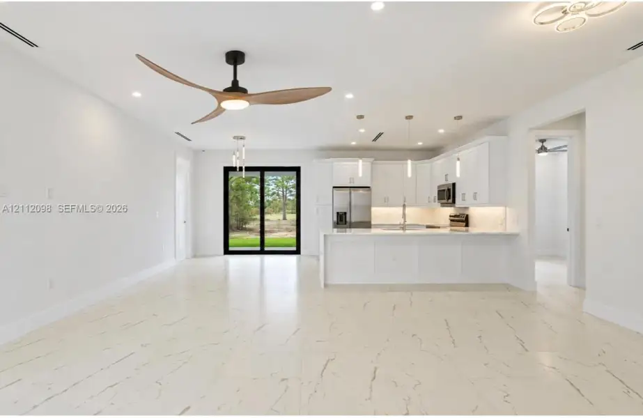 Open-concept area featuring white cabinetry with integrated stainless steel appliances, large kitchen island with light-toned stone-finish countertop, polished porcelain tile flooring, modern three-blade ceiling fan, and sliding glass doors Open-concept area featuring white cabinetry with integrated stainless steel appliances, large kitchen island with light-toned stone-finish countertop, polished porcelain tile flooring, modern three-blade ceiling fan, and sliding glass doors