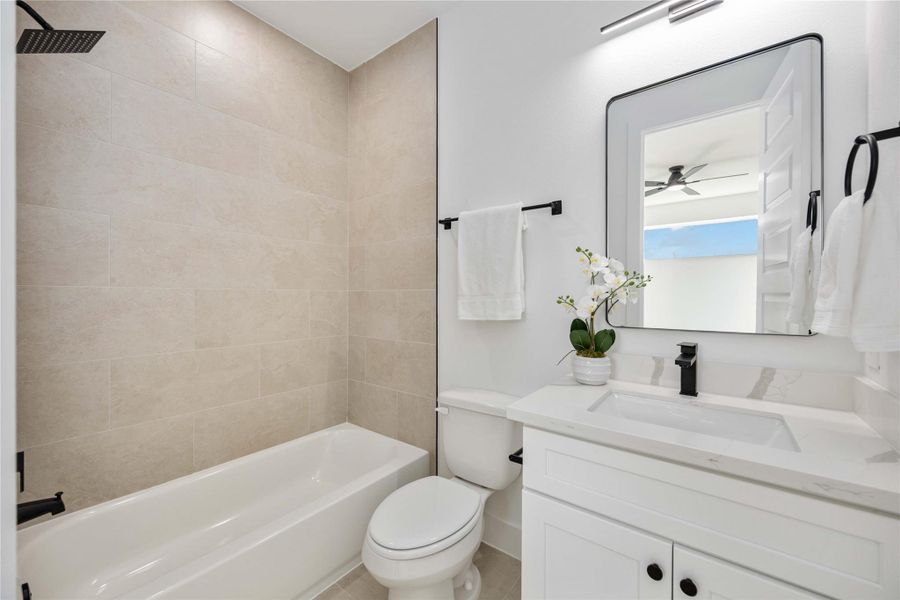 This beautifully updated bathroom features a sleek tub-shower combo with large neutral wall tiles, complemented by a modern white vanity with polished quartz countertops, matte black fixtures, and contemporary lighting that enhances the clean, bright design.