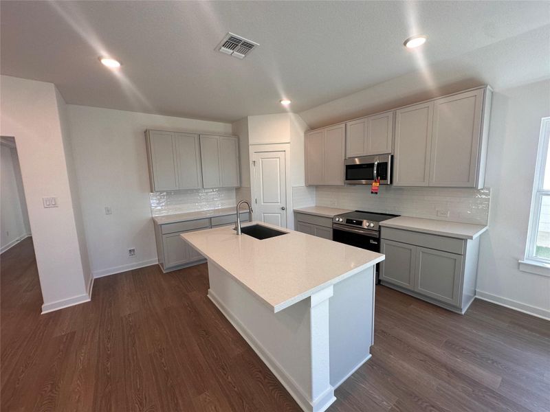 Kitchen featuring gray cabinets, appliances with stainless steel finishes, backsplash, a center island with sink, and light stone counters Kitchen featuring gray cabinets, appliances with stainless steel finishes, backsplash, a center island with sink, and light stone counters
