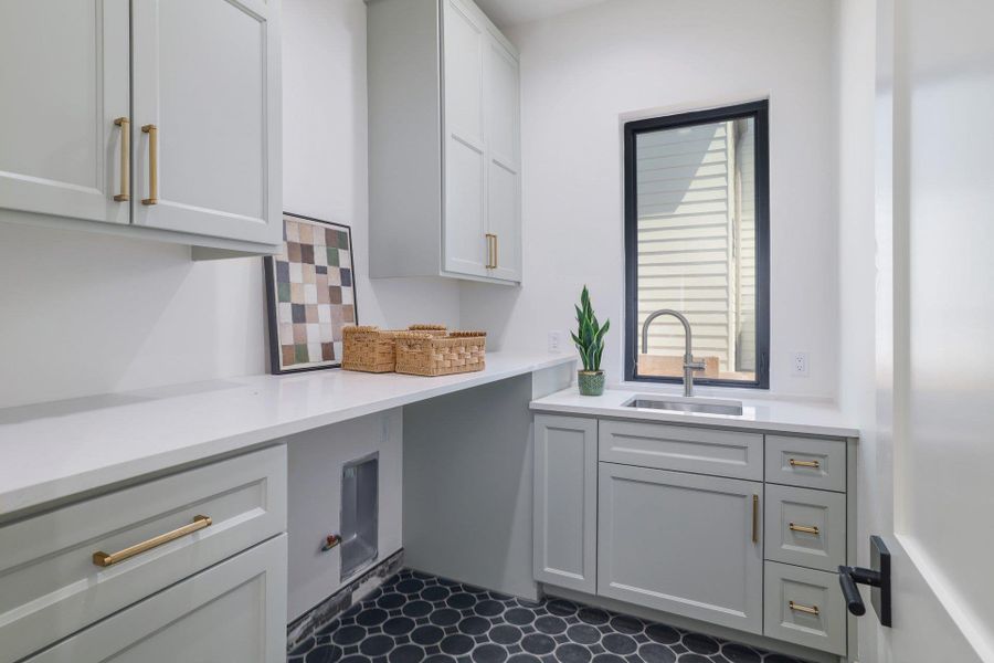 Functional laundry space featuring extensive white countertops, light gray cabinetry with gold-tone hardware, a stainless steel sink with a gooseneck faucet, a black-framed window, and patterned hexagonal floor tiles