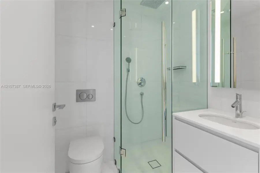 Bathroom featuring a glass-enclosed shower with a rainfall showerhead, wall-mounted dual-flush toilet, white vanity with an integrated sink, polished chrome fixtures, and large-format tile surround