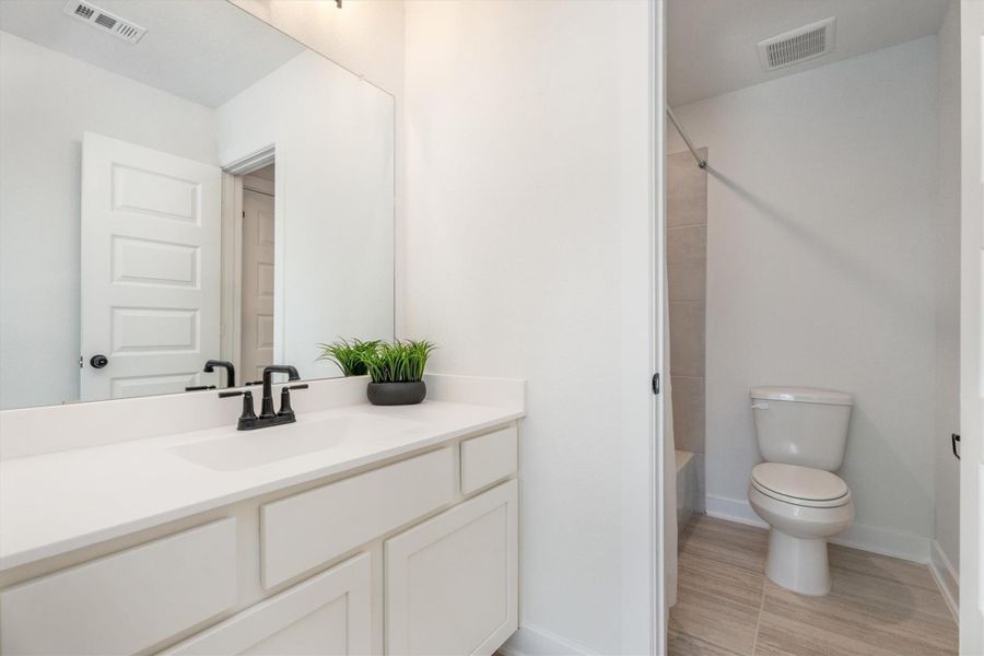This bathroom features a modern, clean design with a large mirror above a white vanity. It includes a toilet and a tub-shower combo, with neutral tile flooring and walls. A small plant adds a touch of greenery.