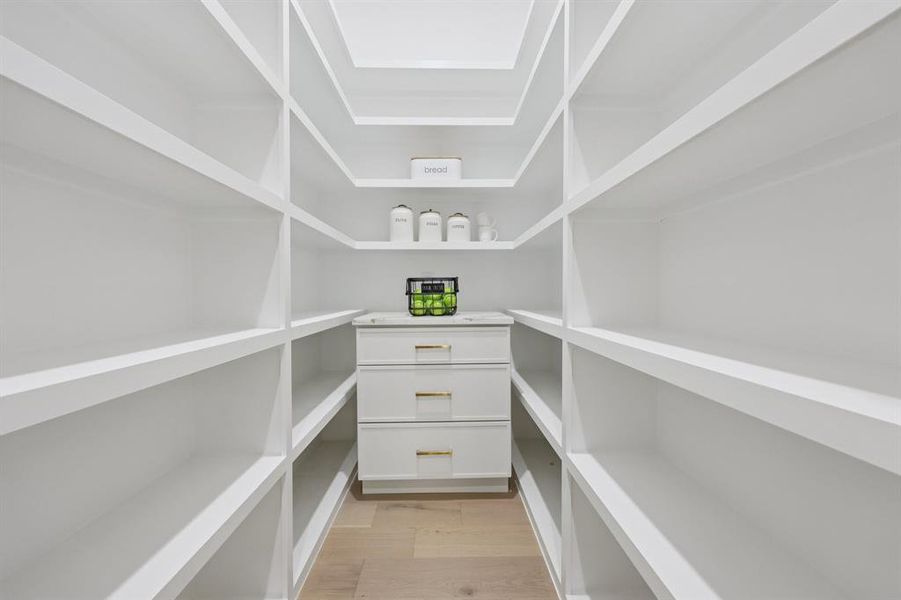 Walk-in pantry featuring extensive white shelving, built-in storage drawers with gold-tone hardware, and light wood-finish flooring