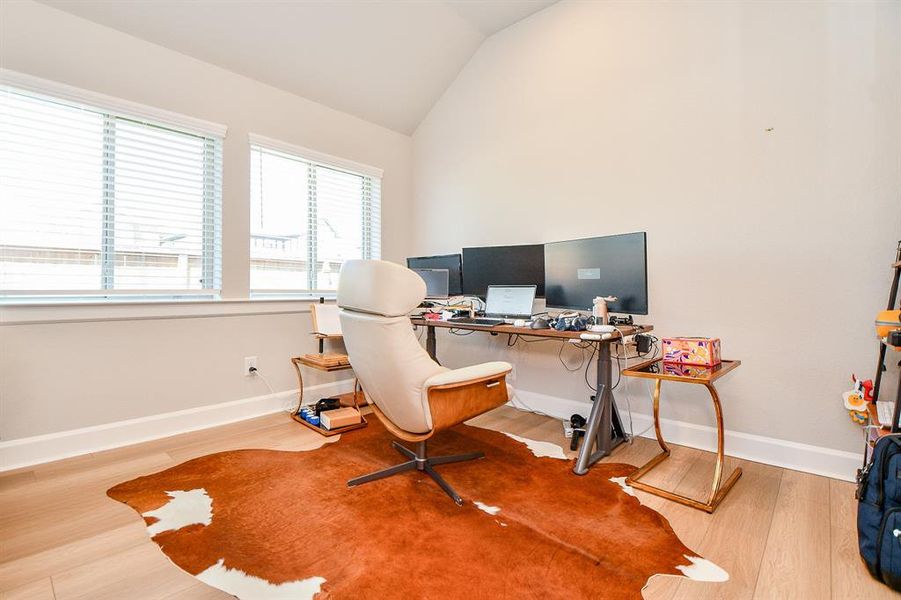 Bright home office with large windows, high ceiling, and wood flooring. Features a modern chair, desk setup with multiple monitors, and a stylish rug. Ideal for productivity and comfort.