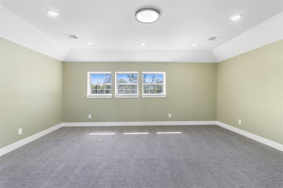 This spacious room features soft green walls, plush gray carpeting, and ample natural light through three large windows. Recessed lighting and a central ceiling fixture provide ample illumination, creating a bright and welcoming atmosphere.