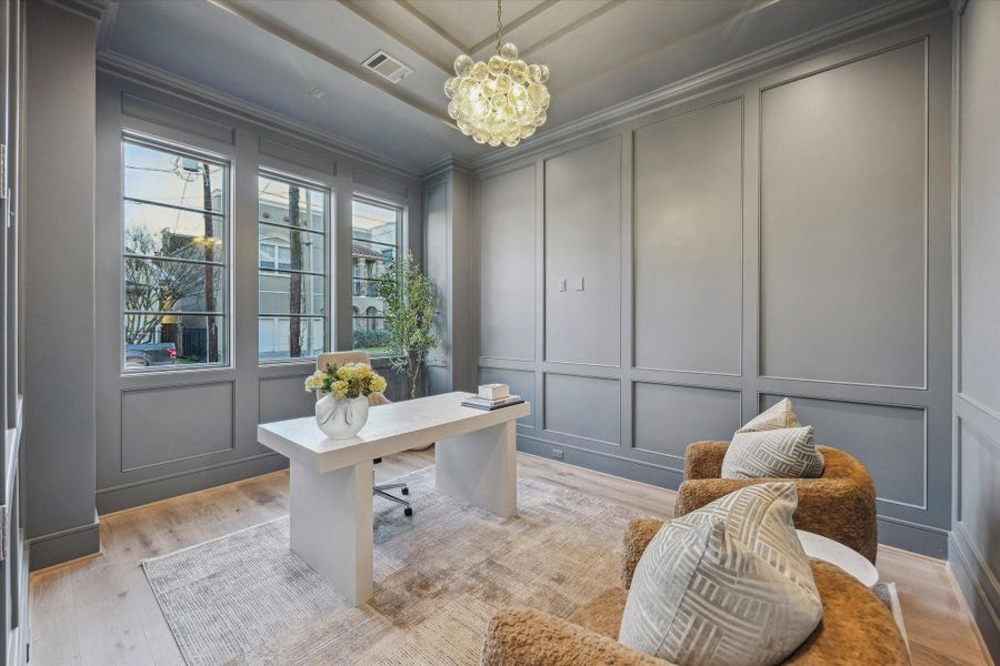 The front office showcases a stepped tray ceiling and wainscoting throughout. The medium gray tones parallel those of the exterior of the home and offer a moody, focused atmosphere. The contemporary bubble light fixture brightens the room at night, while the big windows flood the room with light during the day.