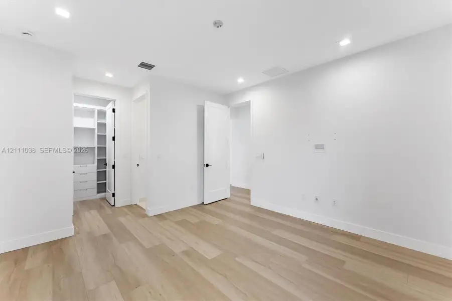 Neutral-toned space featuring wood-finish flooring, recessed lighting, shaker-style doors with matte black hardware, and an integrated walk-in closet with built-in shelving