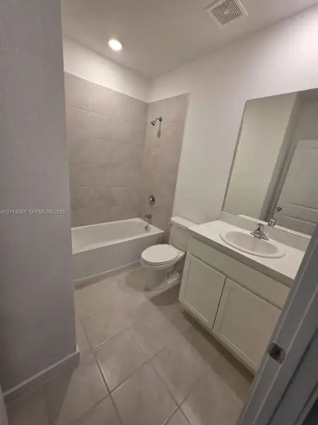 Bathroom featuring neutral tile flooring, a shower-tub combination with full-height wall tiling, a white vanity cabinet, a solid-surface countertop with an integrated basin, and a wall-mounted mirror