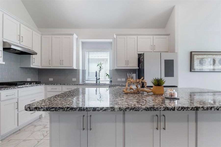 Kitchen featuring dark stone countertops, light marble finish flooring, tasteful backsplash, lofted ceiling, and under cabinet range hood