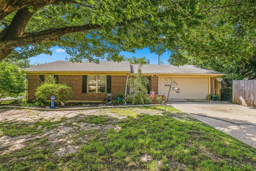 Brick facade home with a two-car garage, mature trees providing shade, a paved driveway, and a fenced side yard Brick facade home with a two-car garage, mature trees providing shade, a paved driveway, and a fenced side yard