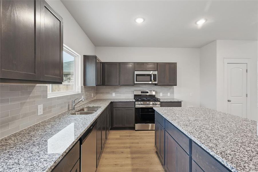 Kitchen with stainless steel appliances, light wood finished floors, light stone counters, dark wood finish cabinetry, and tasteful backsplash