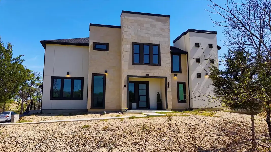 Modern two-story home with a sleek design featuring large windows and a minimalist facade. Set on a slightly elevated plot, surrounded by sparse trees and a clear blue sky, offering a contemporary and spacious feel.