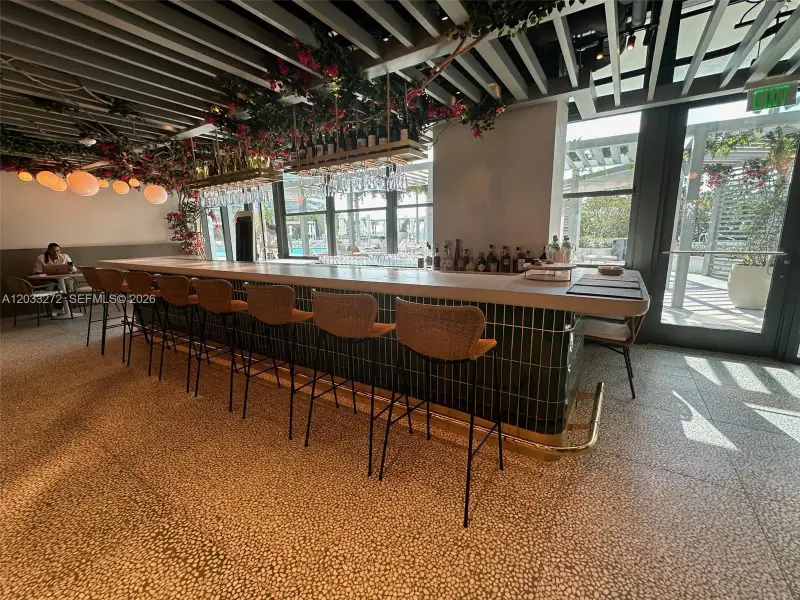 Bar area featuring a polished solid-surface countertop, dark green vertical-tiled base, brass foot rail, and overhead slat ceiling detail with integrated lighting Bar area featuring a polished solid-surface countertop, dark green vertical-tiled base, brass foot rail, and overhead slat ceiling detail with integrated lighting