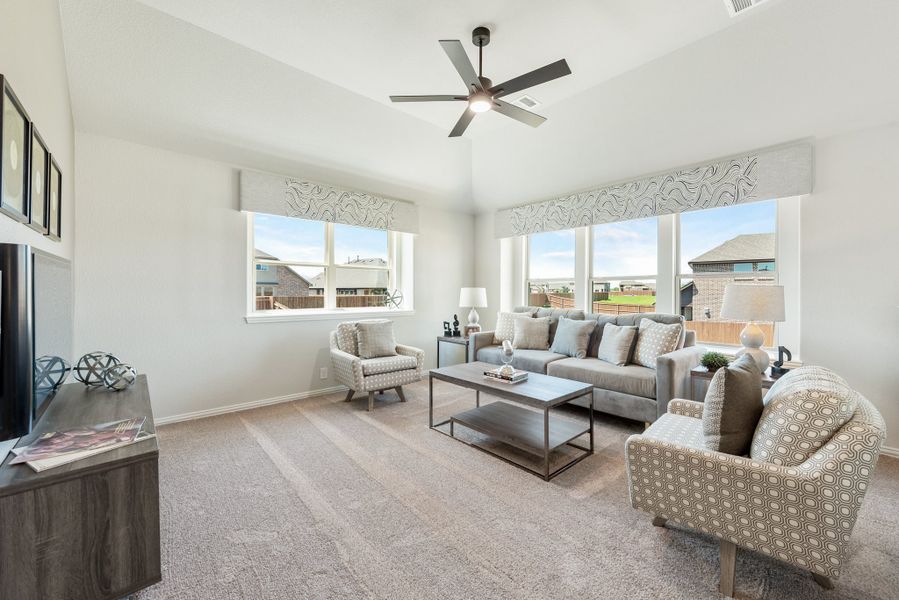 Living room with gray sofa, patterned chairs, ceiling fan, and large windows with neighborhood views
