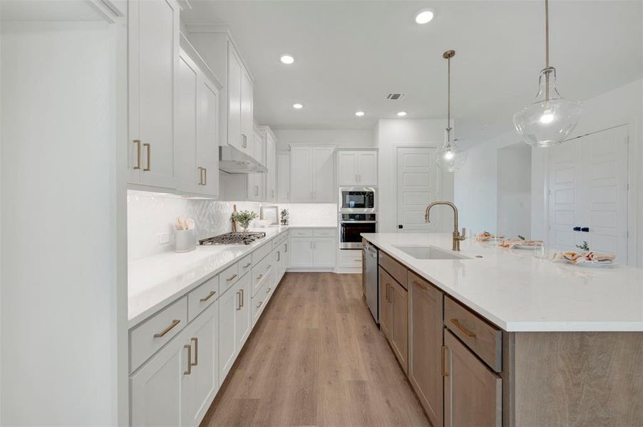 Modern kitchen featuring white shaker cabinetry with gold-tone hardware, a light-toned wood-finish flooring, a central island with an undermount sink and gooseneck faucet, and recessed lighting with two glass pendant fixtures Modern kitchen featuring white shaker cabinetry with gold-tone hardware, a light-toned wood-finish flooring, a central island with an undermount sink and gooseneck faucet, and recessed lighting with two glass pendant fixtures