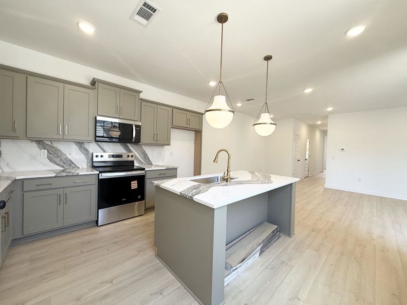 Kitchen featuring stainless steel appliances, gray cabinets, light stone counters, an island with sink, and pendant lighting