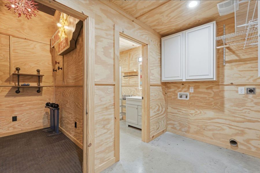 This photo shows a utility room with plywood walls, built-in shelving, and a white cabinet. It features exposed outlets and a space for a washer and dryer. A small adjoining area has additional shelving and lighting in the shop area.