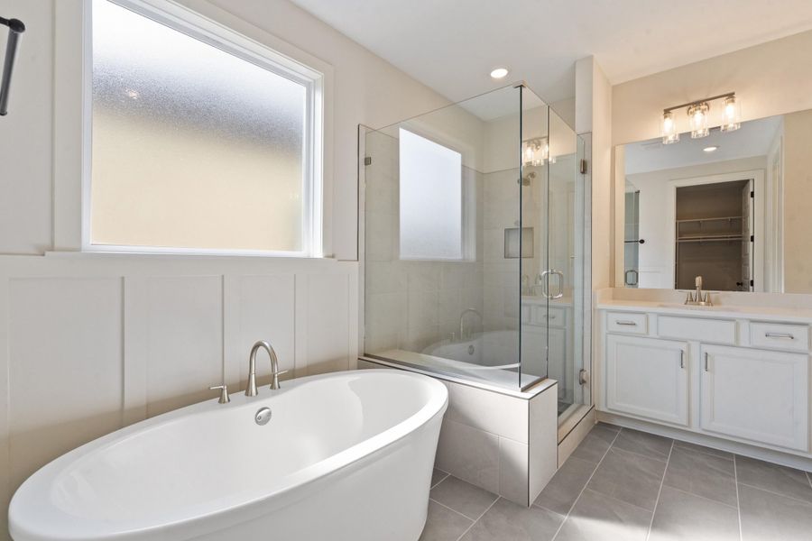 Freestanding soaking tub with brushed nickel fixtures, glass-enclosed shower, white vanity with dual-handle faucet, large vanity mirror, and grey tile flooring Freestanding soaking tub with brushed nickel fixtures, glass-enclosed shower, white vanity with dual-handle faucet, large vanity mirror, and grey tile flooring