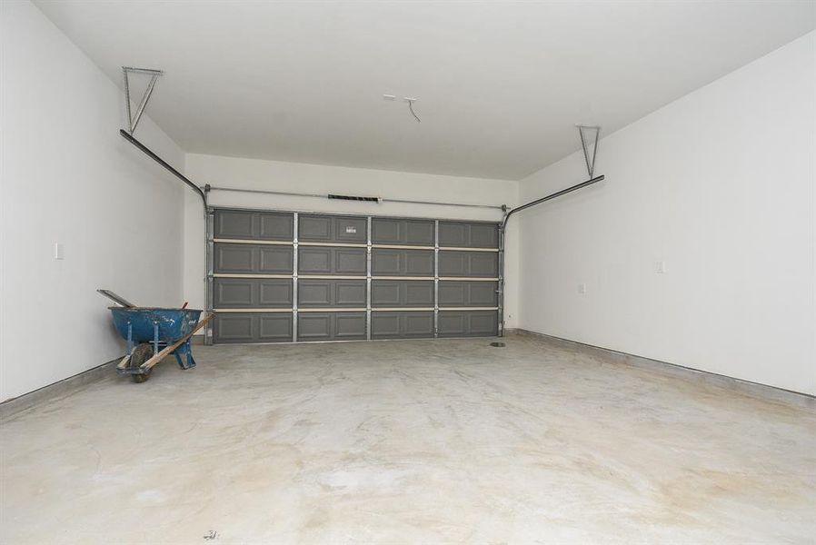 Empty two-car garage with a closed door and a blue wheelbarrow; unfinished, clean concrete floor and white walls, with visible overhead garage door tracks.