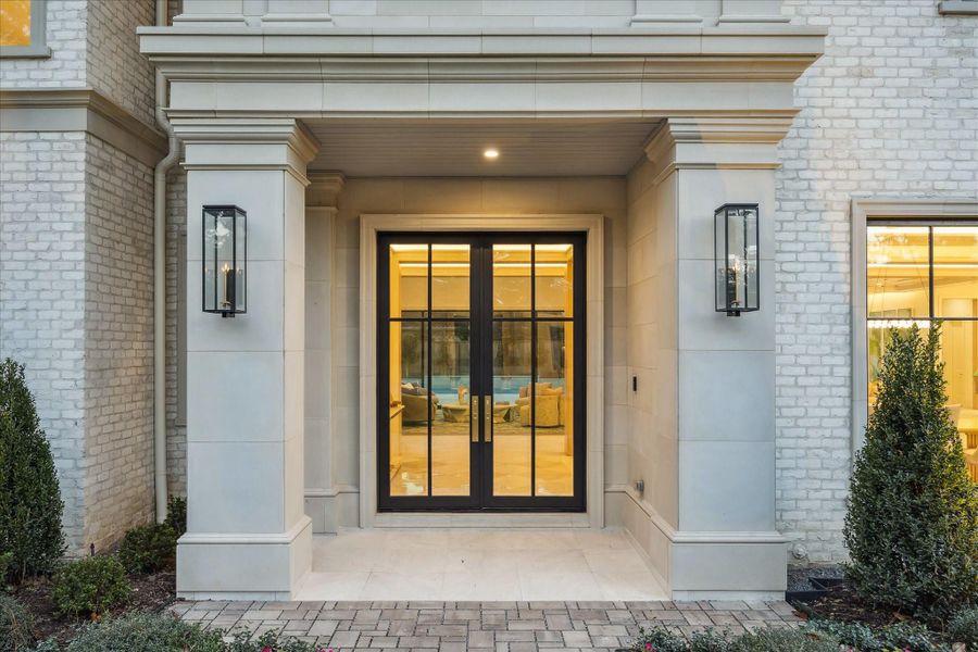 Grand front entry anchored by a steel and glass double door framed by limestone columns and trim detailing. The covered porch includes a clean-lined ceiling with recessed lighting, while modern gas lantern sconces add an elegant touch. Brick façade and neatly manicured landscaping enhance the sophisticated, stately curb appeal.