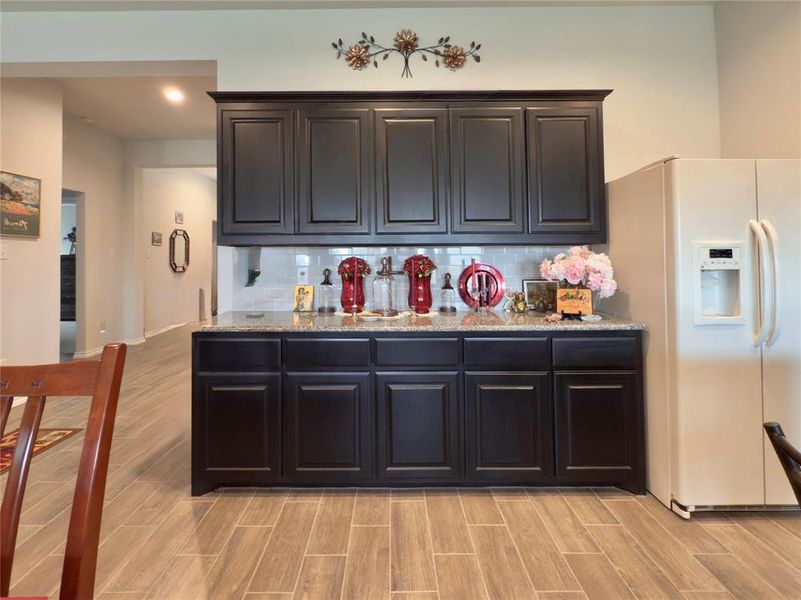 Kitchen featuring white fridge with ice dispenser, light stone countertops, wood tiled floors, tasteful backsplash, and dark cabinetry Kitchen featuring white fridge with ice dispenser, light stone countertops, wood tiled floors, tasteful backsplash, and dark cabinetry