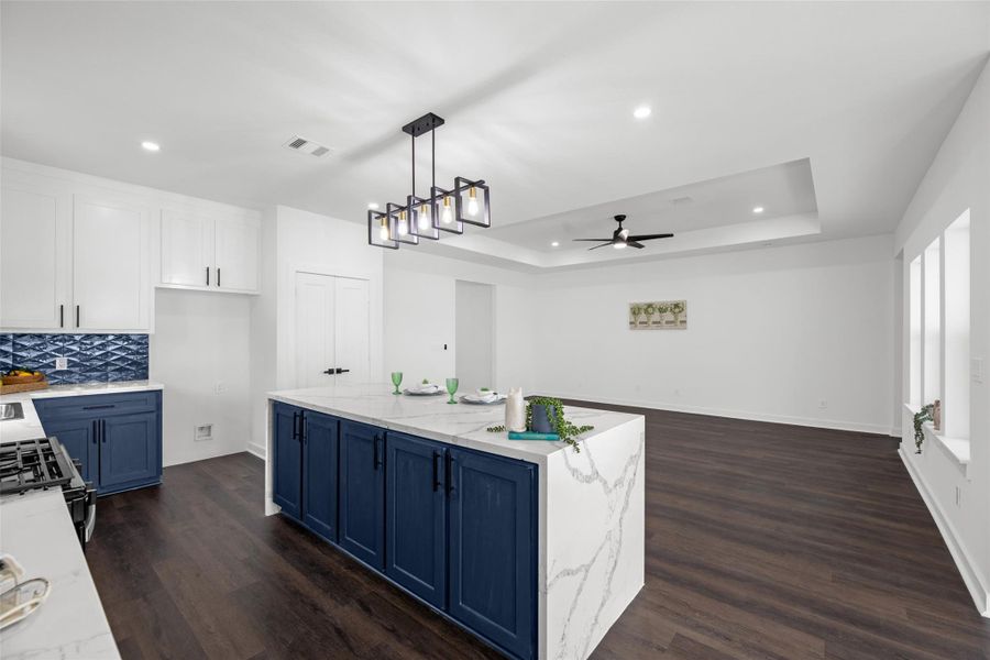 Large island with blue cabinetry, a sleek marble countertop, and a stylish light fixture above. White cabinets and a striking blue tile backsplash provide a fresh contrast, while the spacious living area includes recessed lighting and a ceiling fan, creating a bright and airy space.