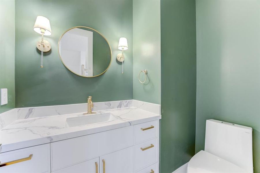 The beautifully painted green half bathroom  has a gorgeous circular mirror, gold hardware, quartz countertops, and chic white cabinetry.