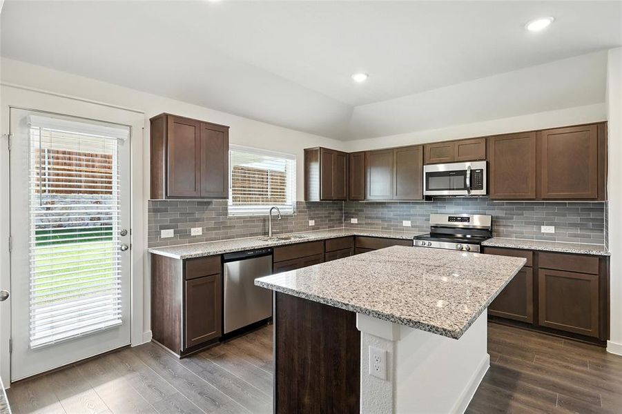 Kitchen with dark wood-style floors, stainless steel appliances, a center island, tasteful backsplash, and light stone counters Kitchen with dark wood-style floors, stainless steel appliances, a center island, tasteful backsplash, and light stone counters