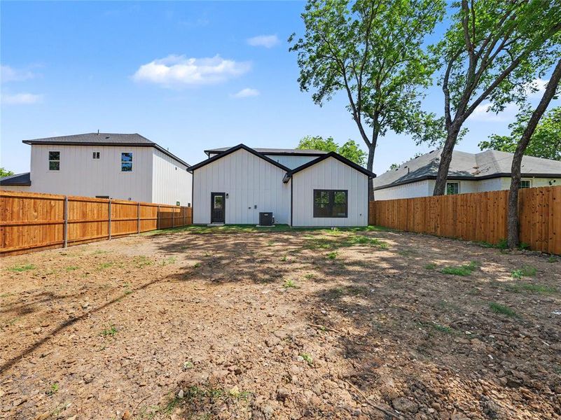 Expansive backyard featuring a new privacy fence, a new construction home with white vertical siding and black trim, a rear entry door, and large mature trees Expansive backyard featuring a new privacy fence, a new construction home with white vertical siding and black trim, a rear entry door, and large mature trees