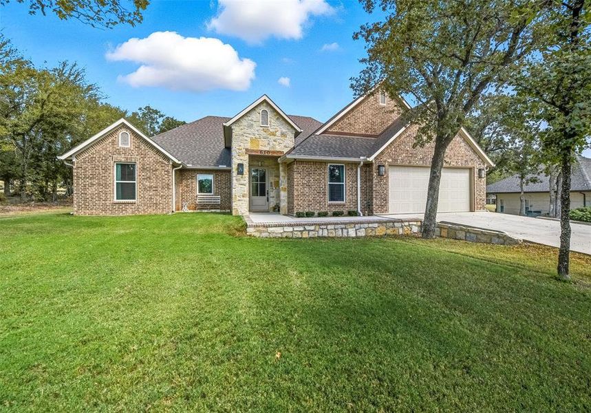 View of front facade with a shingled roof, a front yard, concrete driveway, stone siding, and brick siding View of front facade with a shingled roof, a front yard, concrete driveway, stone siding, and brick siding